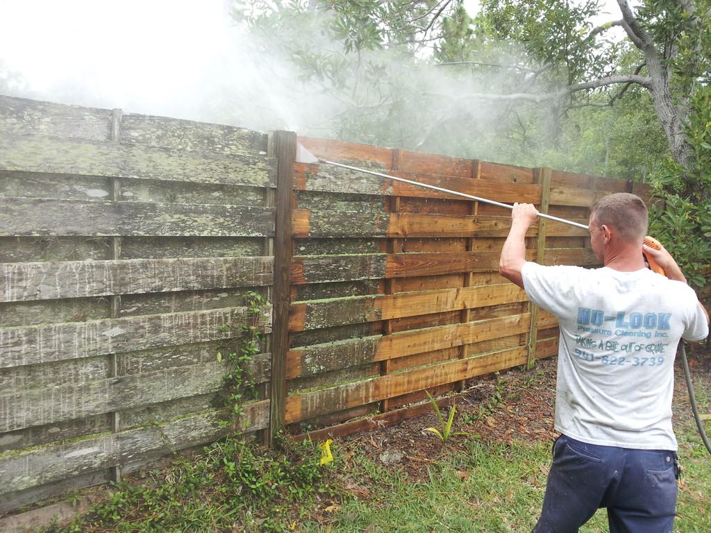 A Nu-Look Pressure Cleaning crew member washing a weathered horizontal wood fence. The cleaned boards on the right are warm brown; the section on the left is still grey and covered in moss.