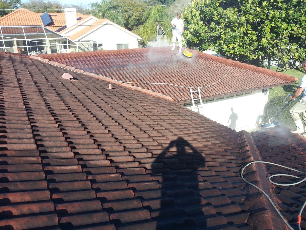 Two Nu-Look crew members cleaning a terracotta barrel tile roof on a Florida home with rotary surface cleaners; the near section of the roof is still dark with algae and mildew.