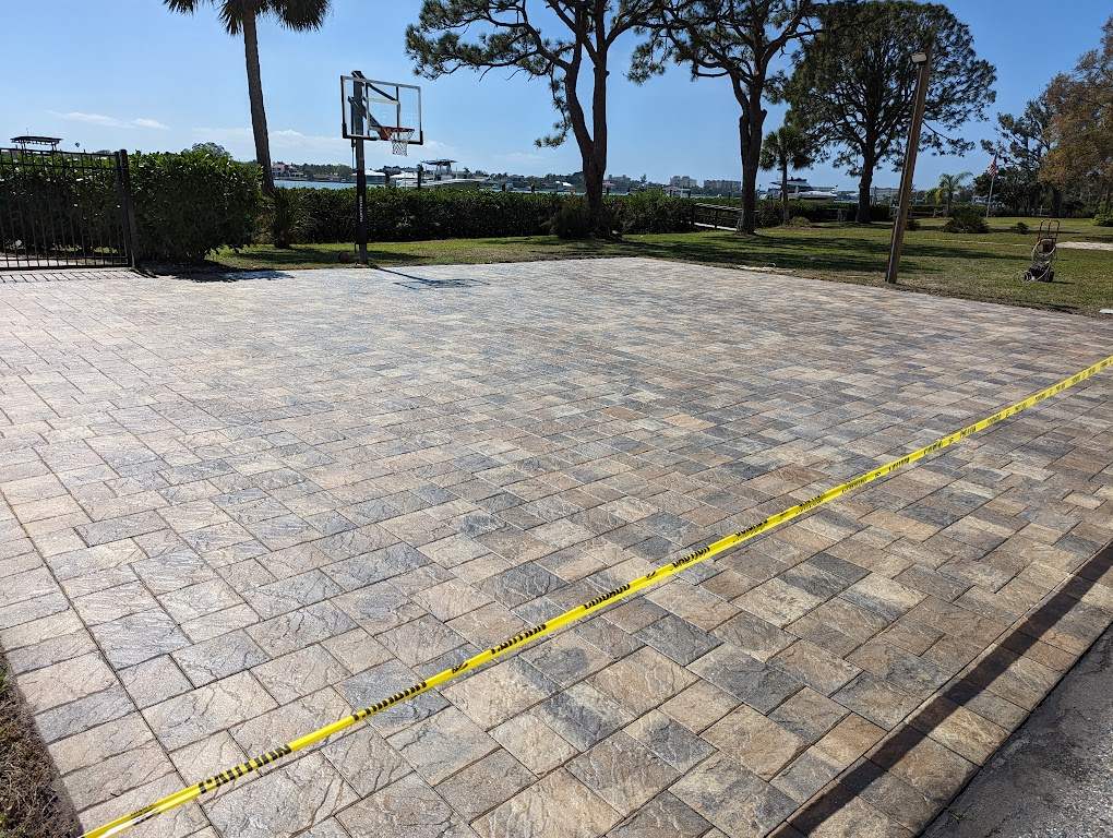 Large paver sport court beside the water, freshly cleaned and sealed, with caution tape across it while the sealer cures.